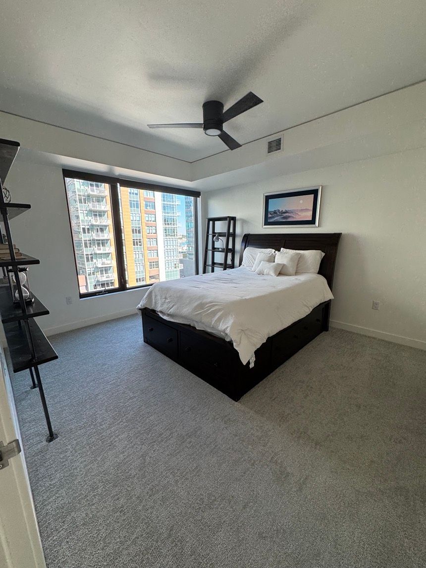 Bedroom with bed, shelves, window, ceiling fan, and city view. Gray carpet and white walls.