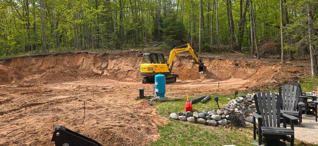 Yellow excavator in a dirt pit, excavating near trees and lawn chairs.
