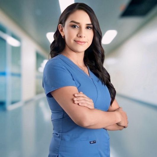 Una mujer con una blusa azul está parada en el pasillo de un hospital con los brazos cruzados.