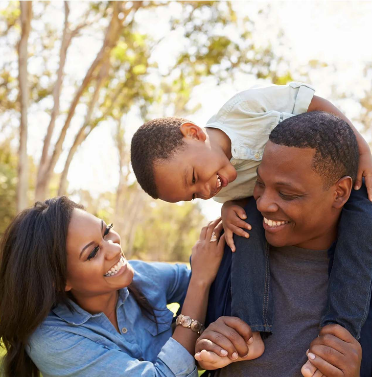 Smiling family outdoors, with a child leaning over a father's shoulders beside a mother in a blue shirt