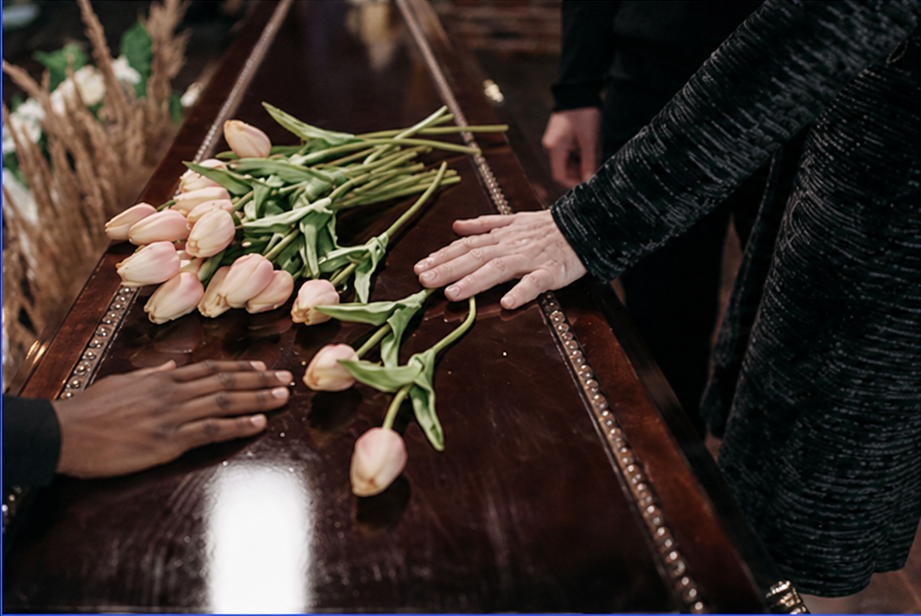 Hands place pale pink tulips on a polished dark wooden casket at a funeral viewing