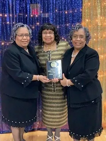 Three women in formal outfits holding a framed plaque, standing before a sparkling backdrop.
