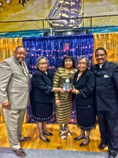 Five people in formal attire pose on a decorated stage, holding a trophy together.