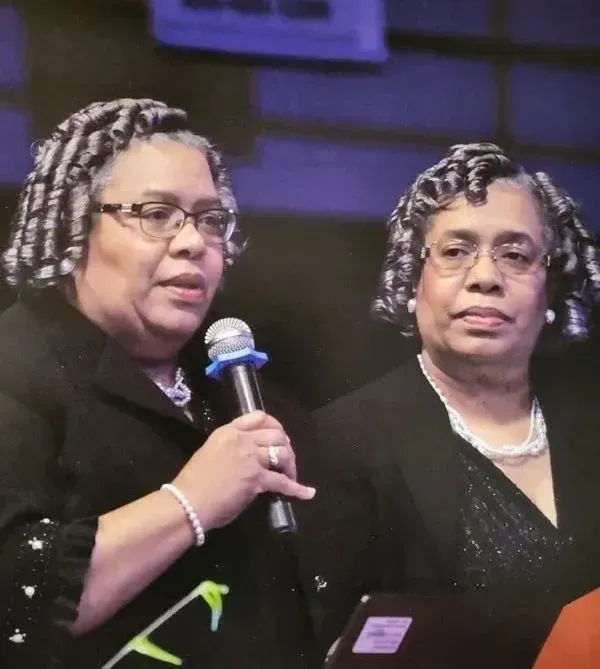 Two women on stage in black outfits, one speaking into a microphone under purple lighting.