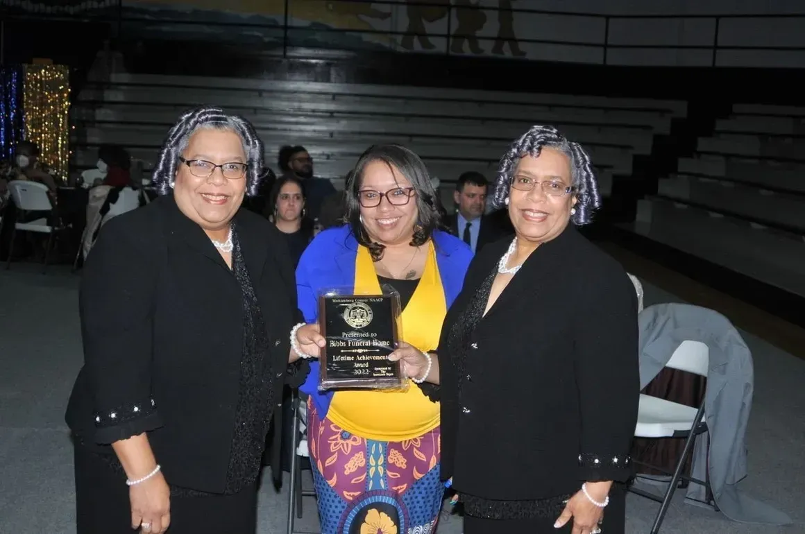 Three women posing indoors, holding an award plaque at an event