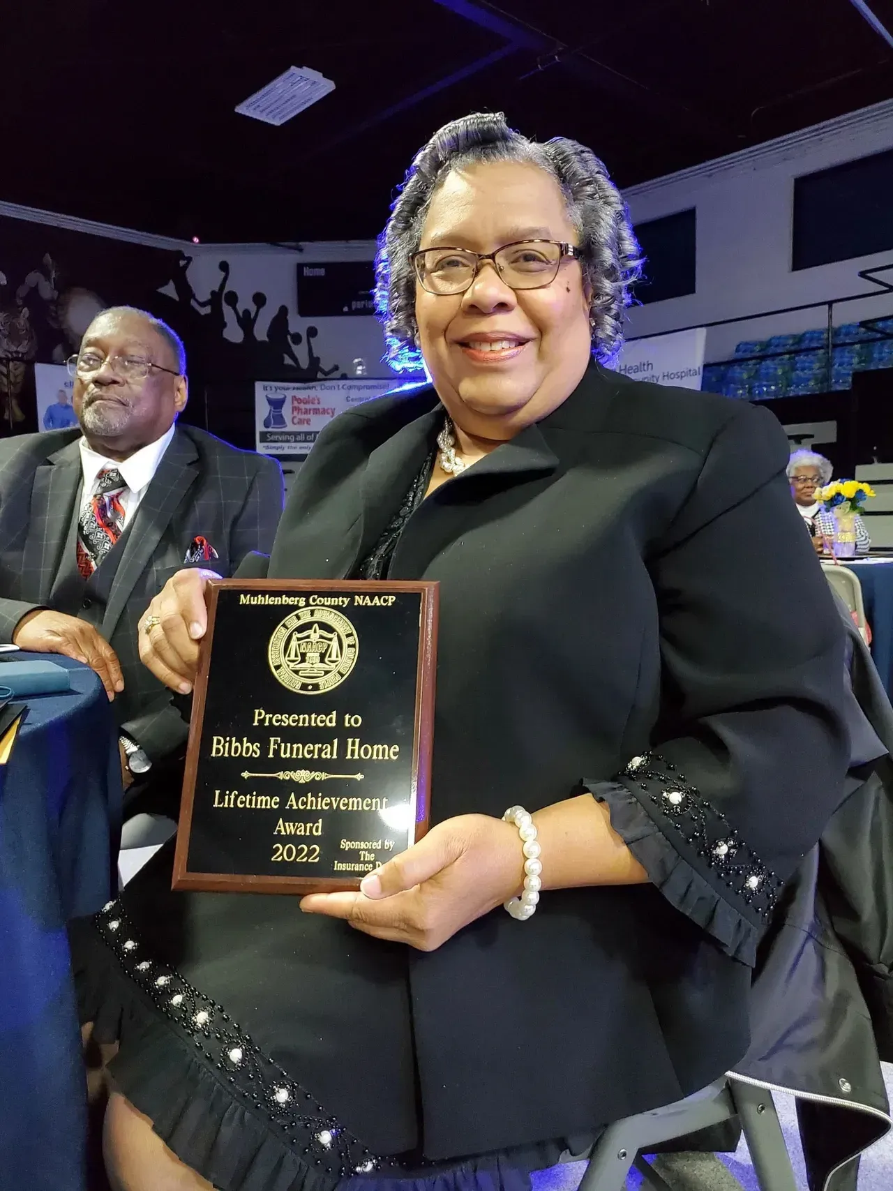 Woman holding award plaque on stage, wearing black outfit with sparkling trim and smiling at ceremony