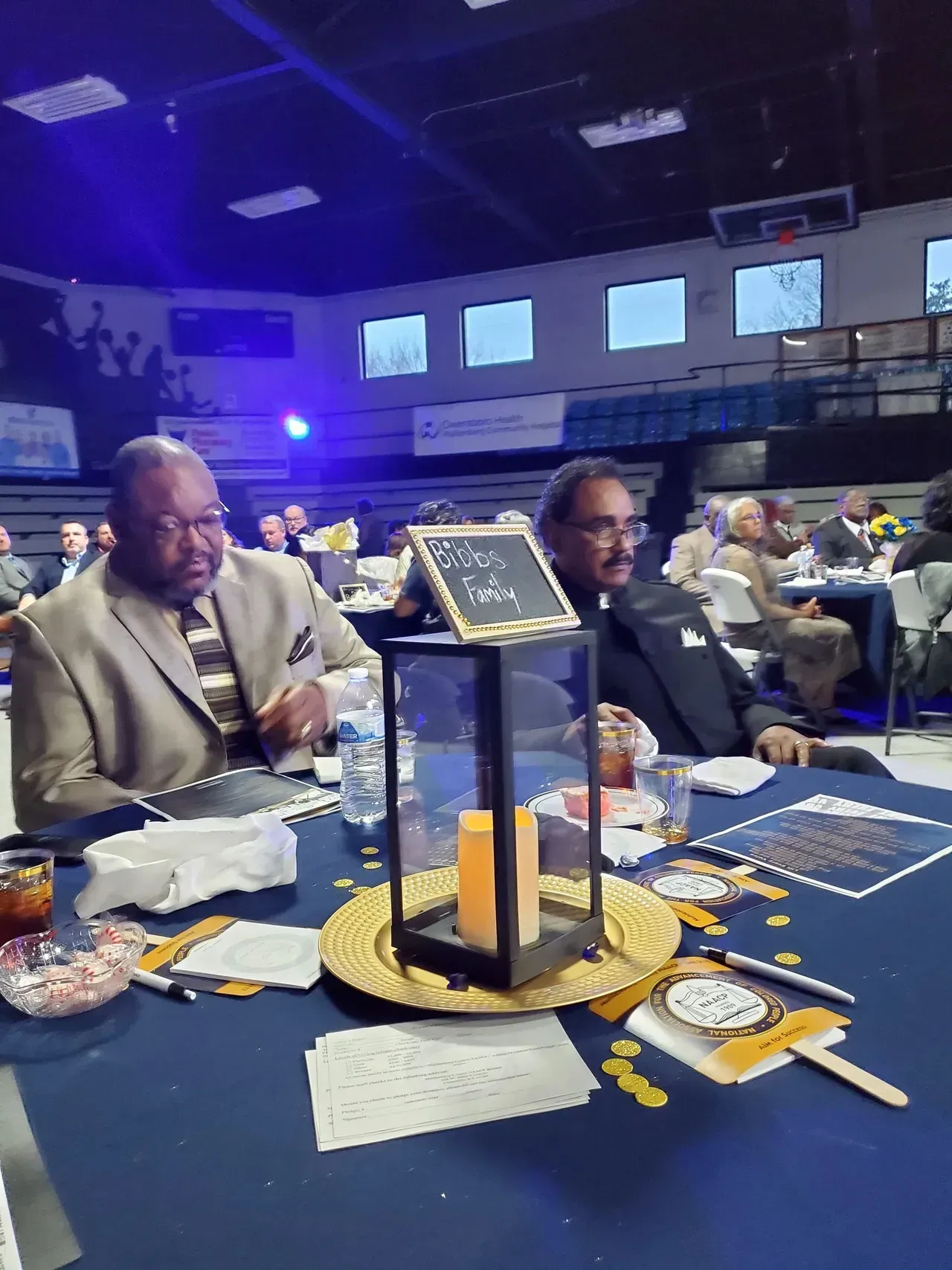 People seated at round banquet tables in a dimly lit event hall with blue lighting and a centerpiece candle arrangement