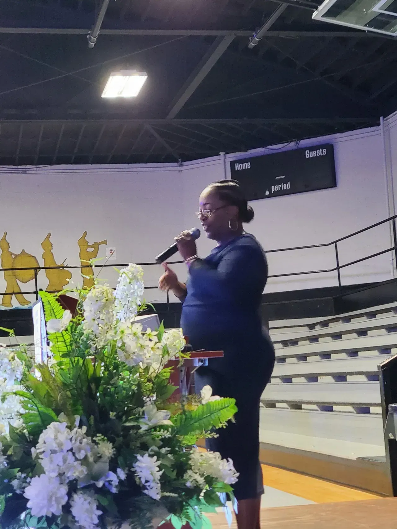 Person speaking into a microphone beside a flower arrangement in a gymnasium or auditorium