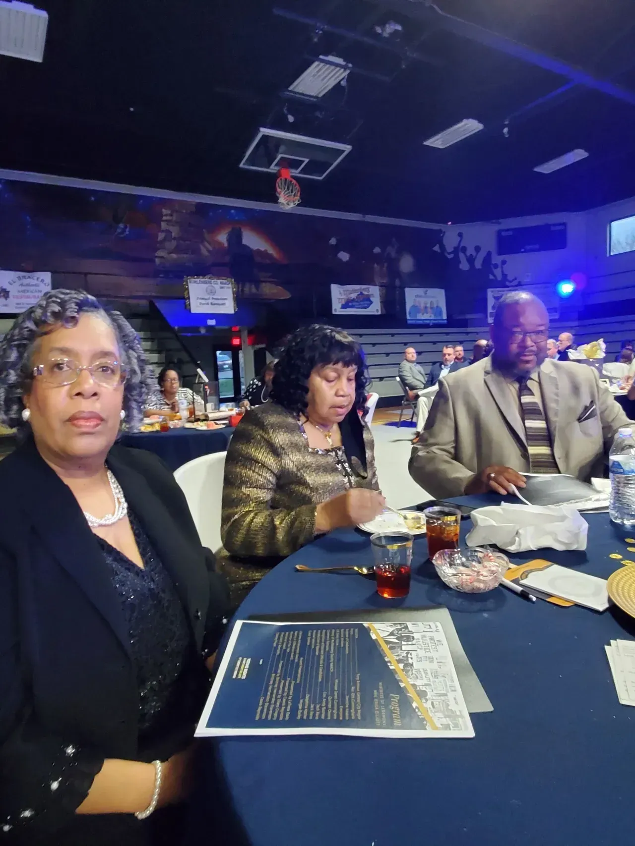Three people seated at a table in a dimly lit event venue, with drinks and menus in front of them.