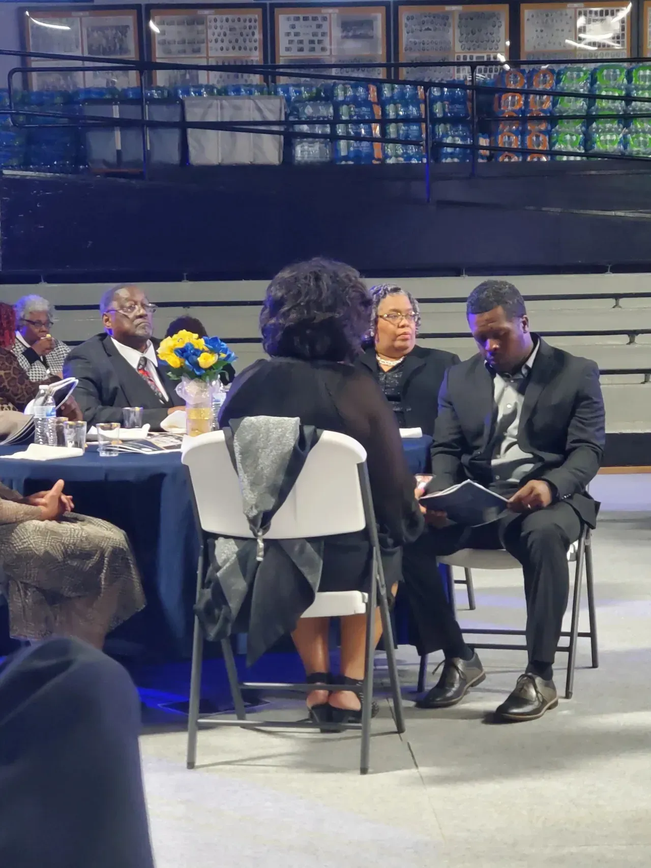 Three people seated and conversing at a formal indoor event, with a stage and tables in the background.
