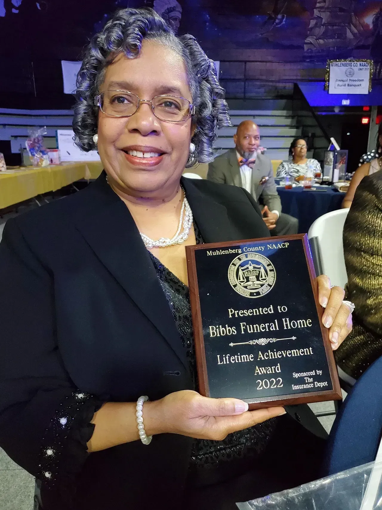 Woman in black blazer holding an award plaque at a banquet event, smiling at the camera.