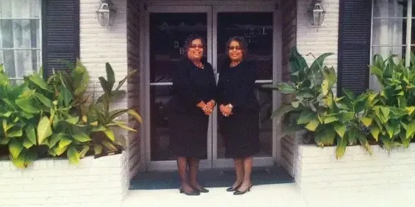 Two women standing on a porch in front of a house, wearing dark outfits and smiling