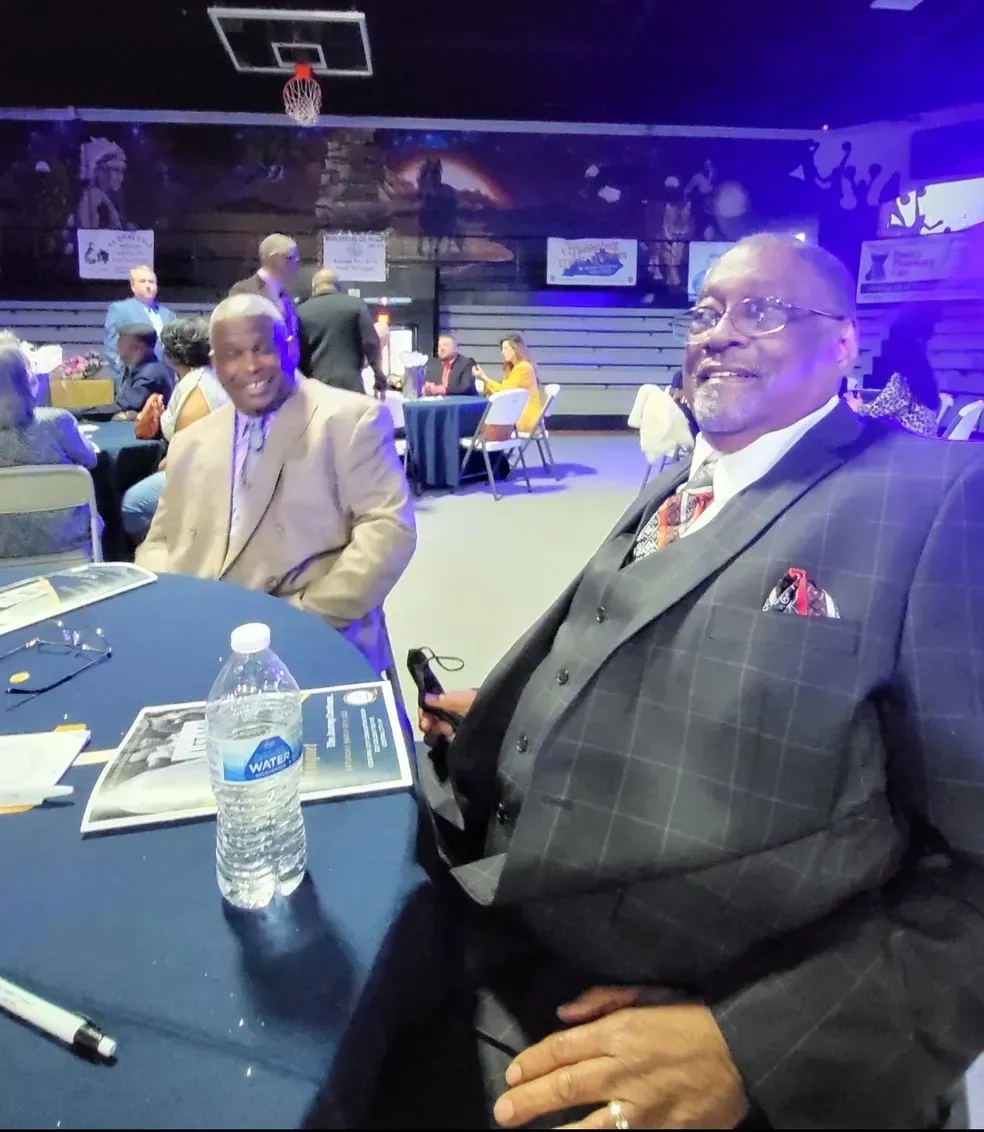 Two men seated at a table in a dim event venue with a basketball hoop in the background