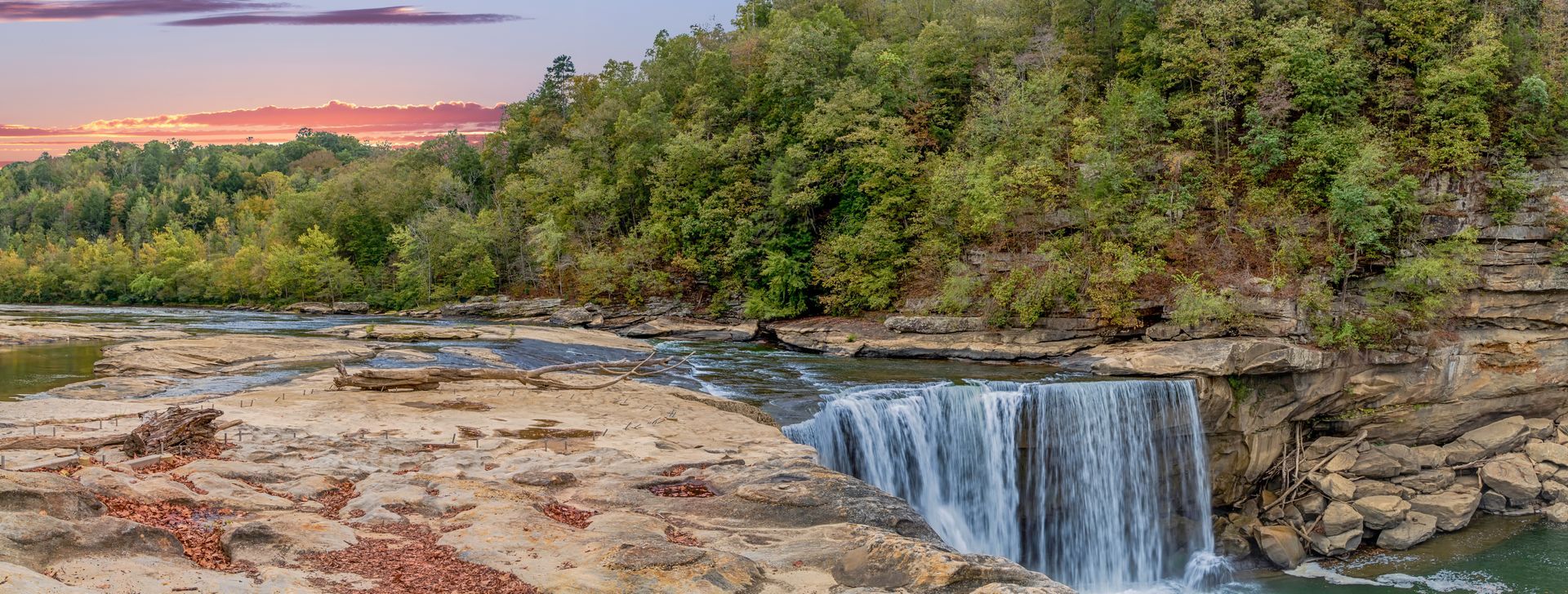 Rocky waterfall at sunset with a pink sky and green trees along the riverbank