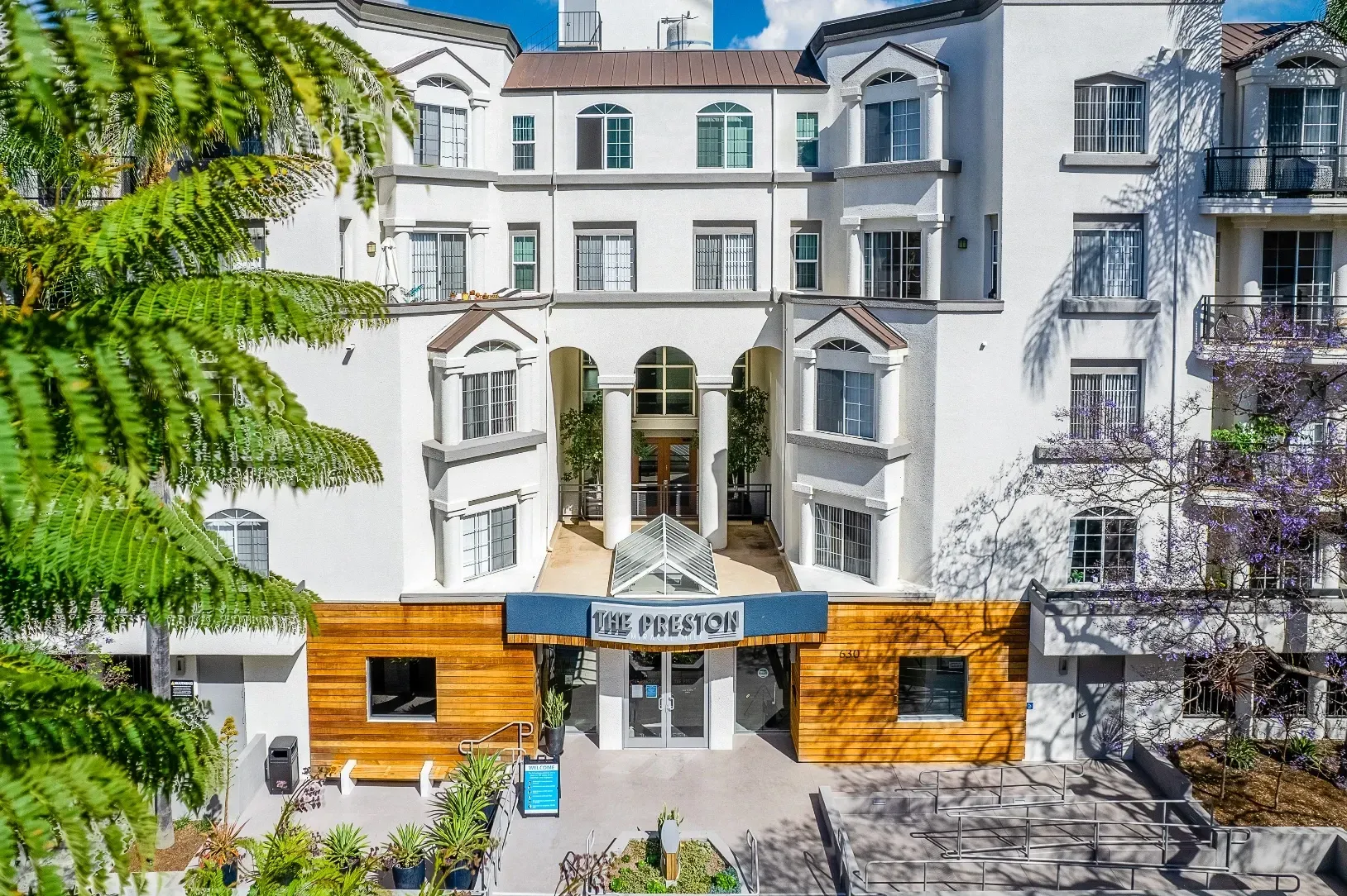 The Preston apartment building entrance and facade with trees and purple flowers in Los Angeles, CA.