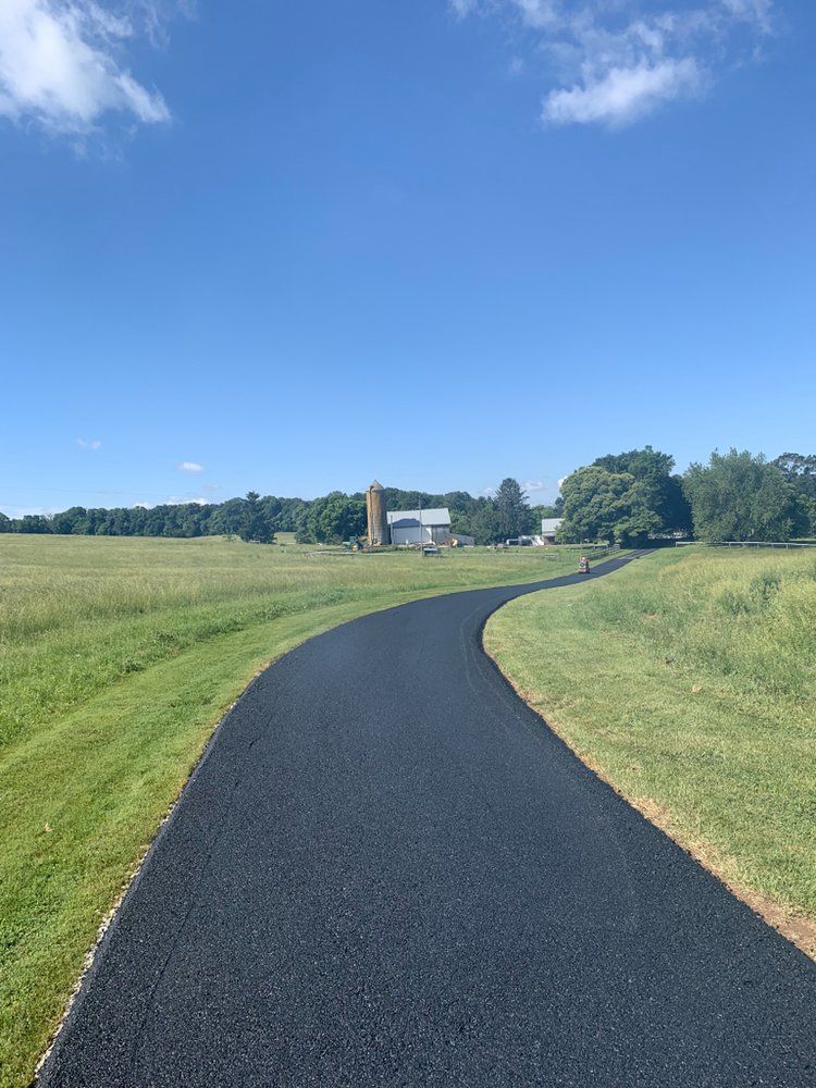 Winding black paved path through green fields under a blue sky, leading to distant trees and a house