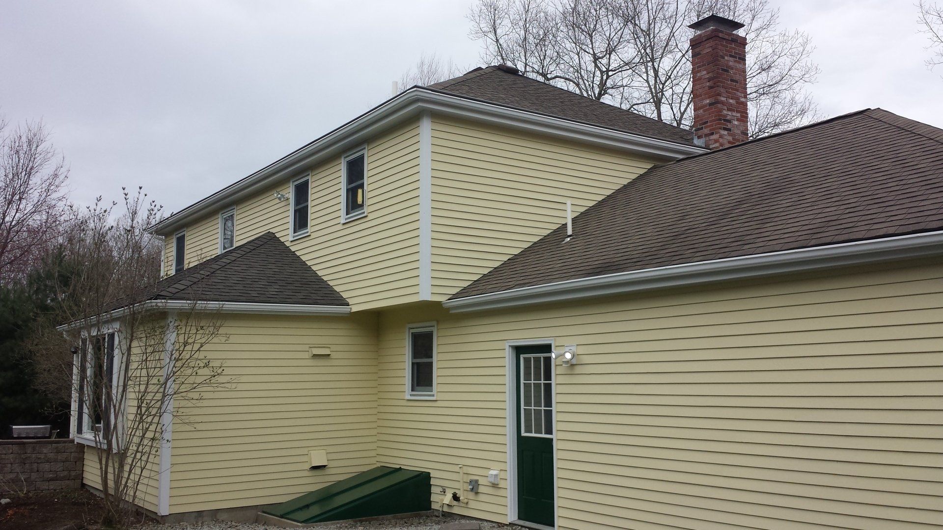 Yellow house with brown roof and brick chimney against an overcast sky.