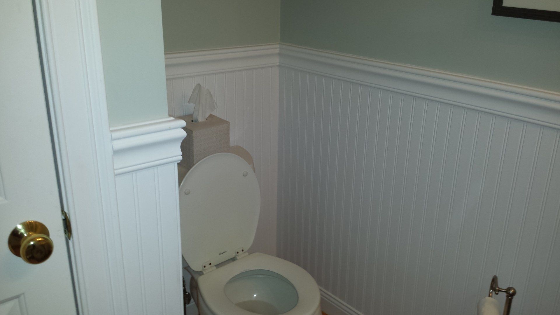 A small bathroom with a toilet and white wainscoting on walls, next to a partially open door.
