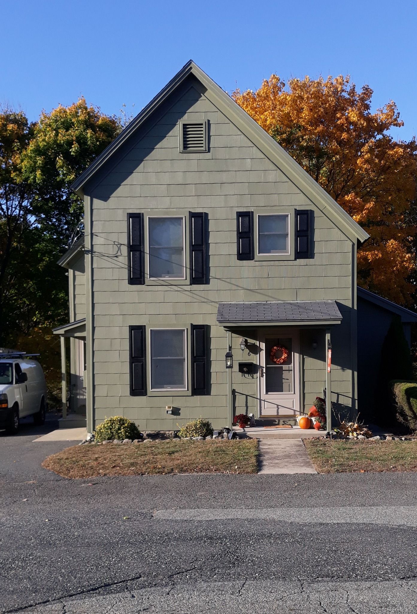 Two-story green house with black shutters, front door, and autumn trees in the background.
