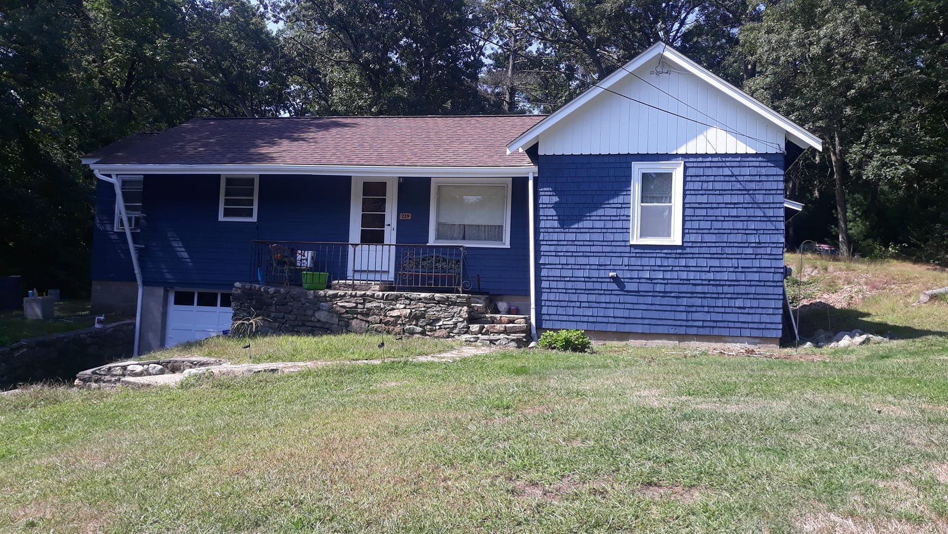 Blue house with white trim, a small front porch, and garage, set against trees and a grassy yard.