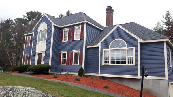 Two-story blue house with white trim, red shutters, and a brick chimney, on a sloped yard with reddish mulch.