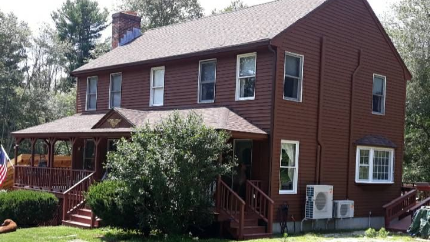 Brown two-story house with a porch and steps, surrounded by green grass and trees.