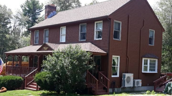 Two-story brown house with a porch. Green lawn and trees surround the home.
