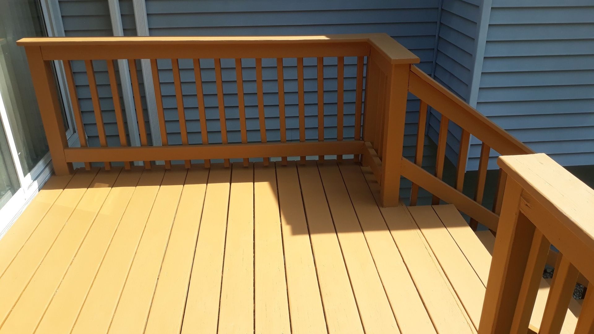 Wooden deck with railing, tan boards, and brown railing, next to a blue wall.