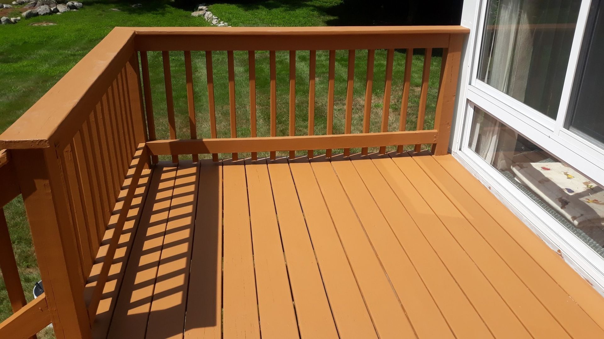 Wooden deck with railing, painted orange-brown, next to a window; grass visible in the background.
