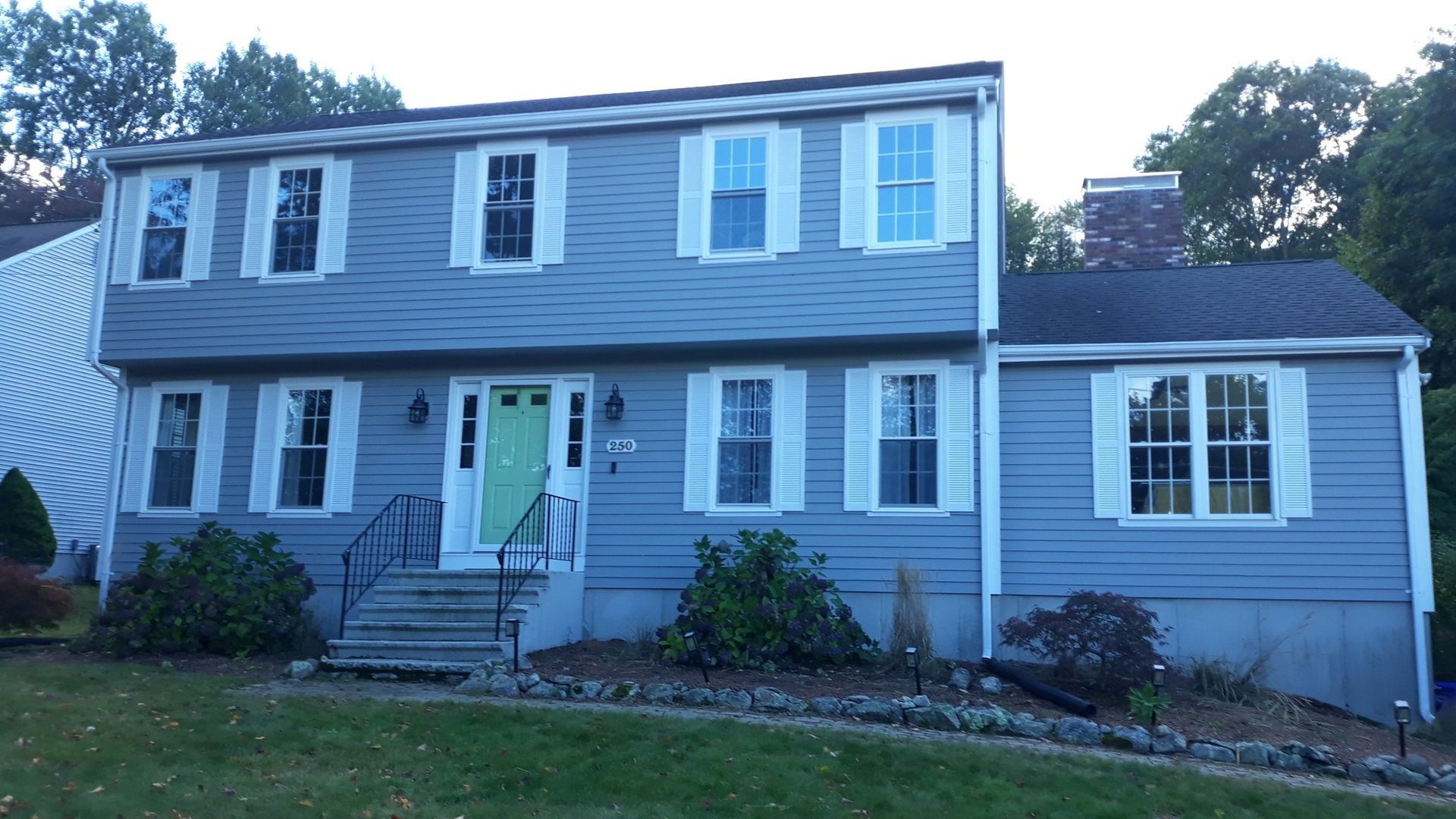 Two-story blue house with white shutters, green door, and stone steps leading to the entrance.