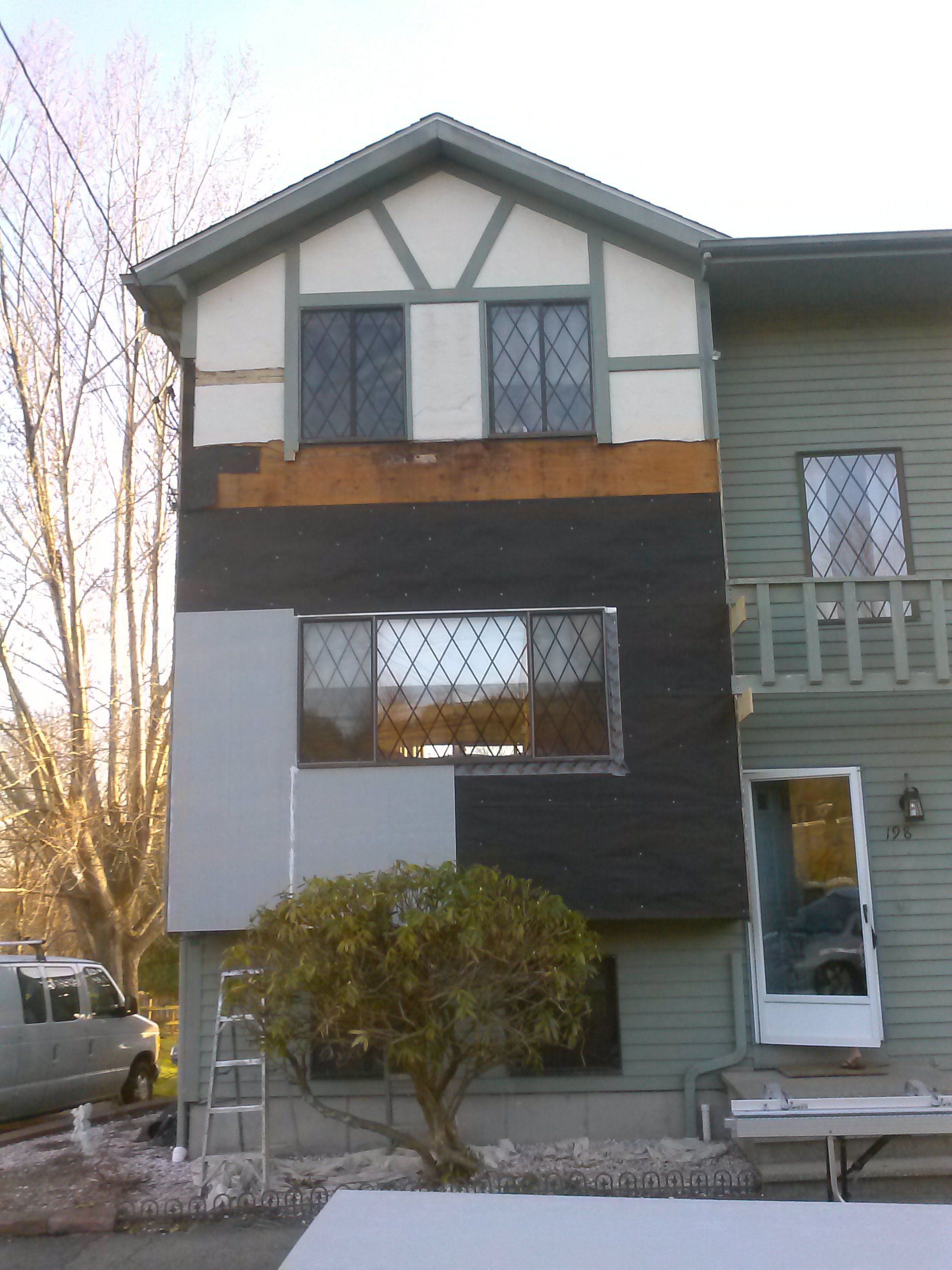 Exterior of a building under construction, showing different siding types and a small tree in front.