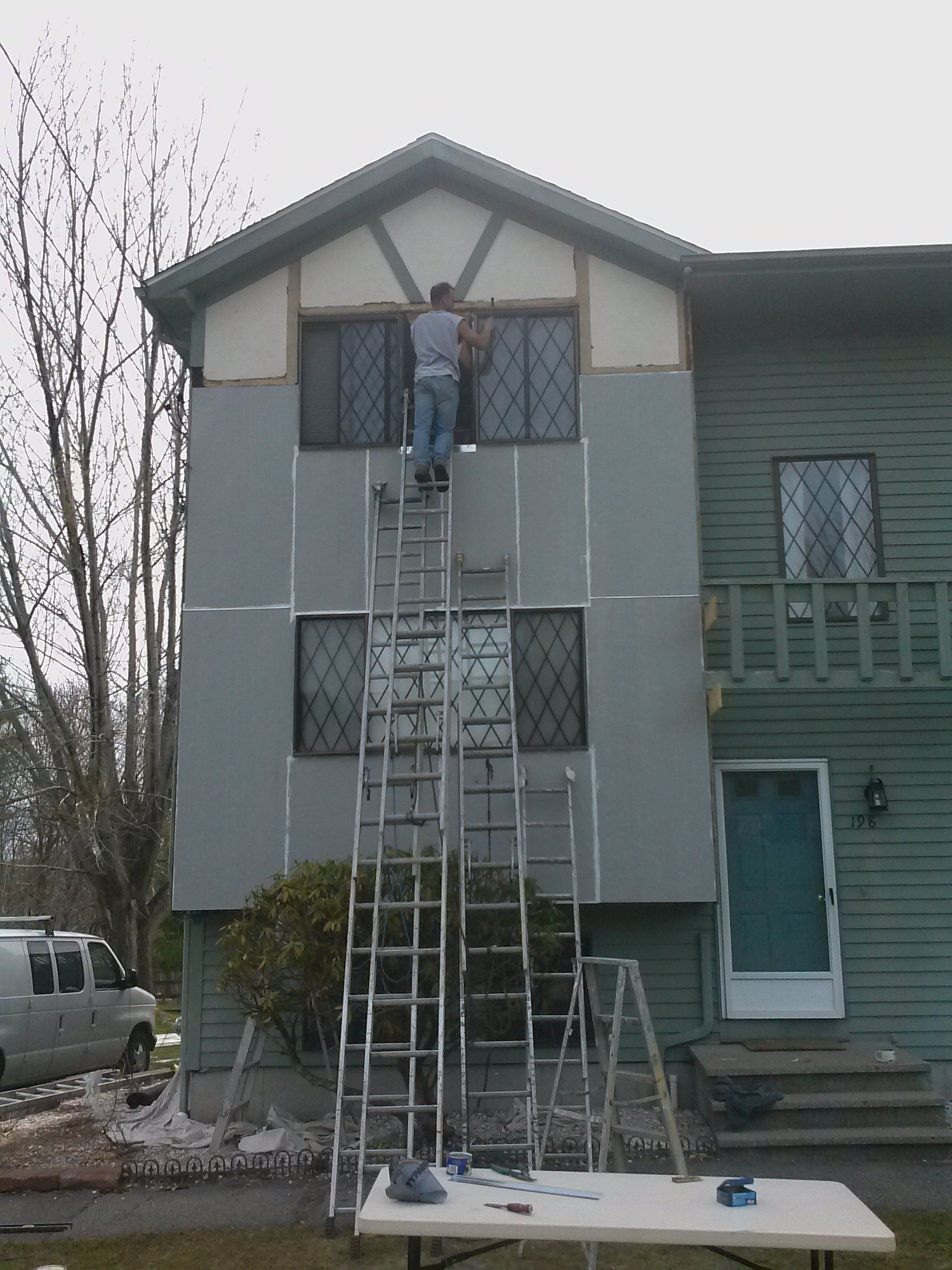 Person on tall ladder working on the side of a two-story house. Gray siding, windows, and tools present.