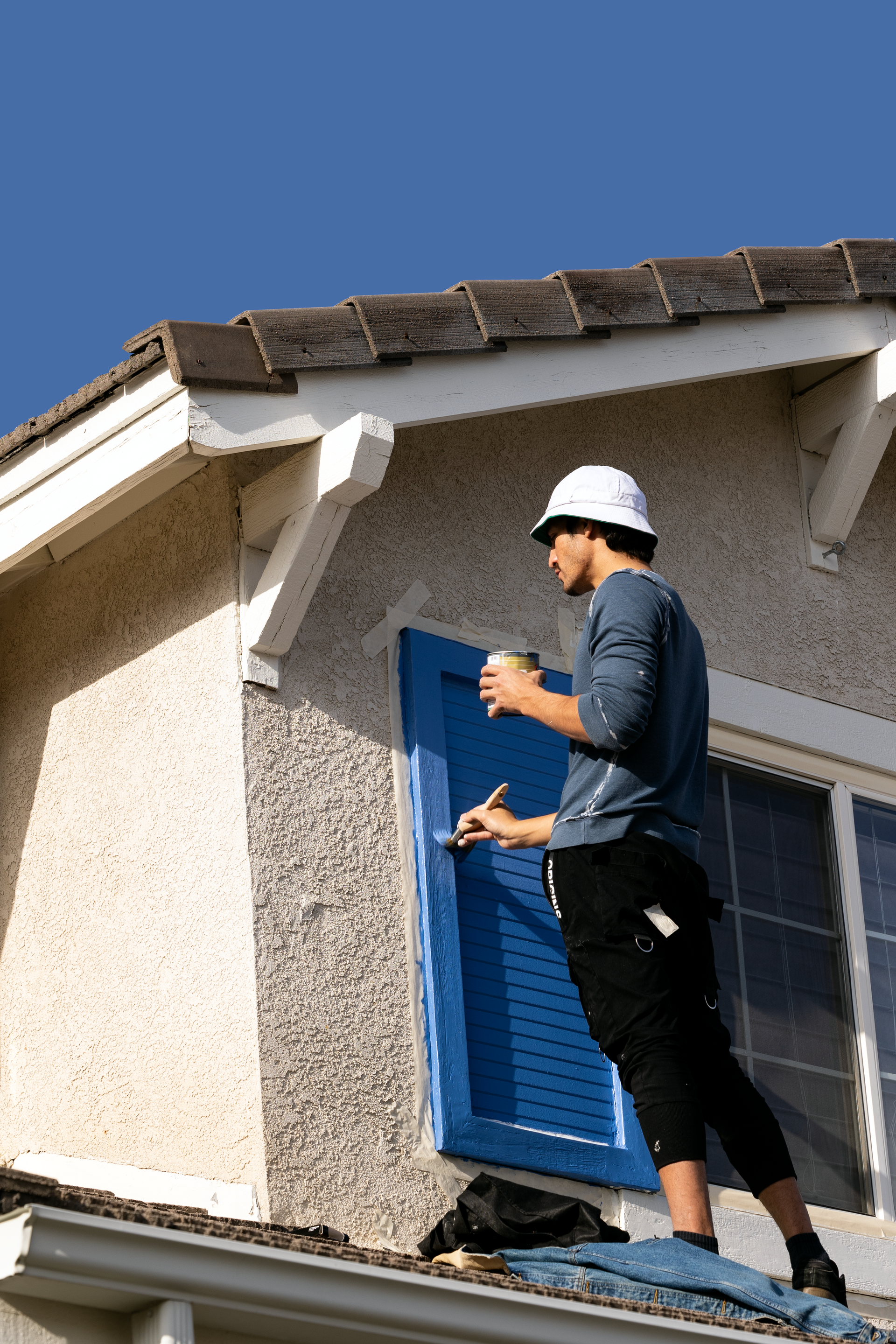 Worker on a roof painting blue shutters on a stucco house.