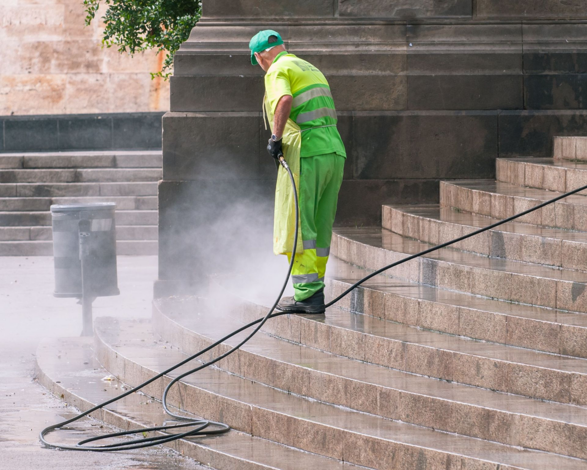 Worker in green safety gear pressure-washing stone steps outdoors, with hose and water spray.