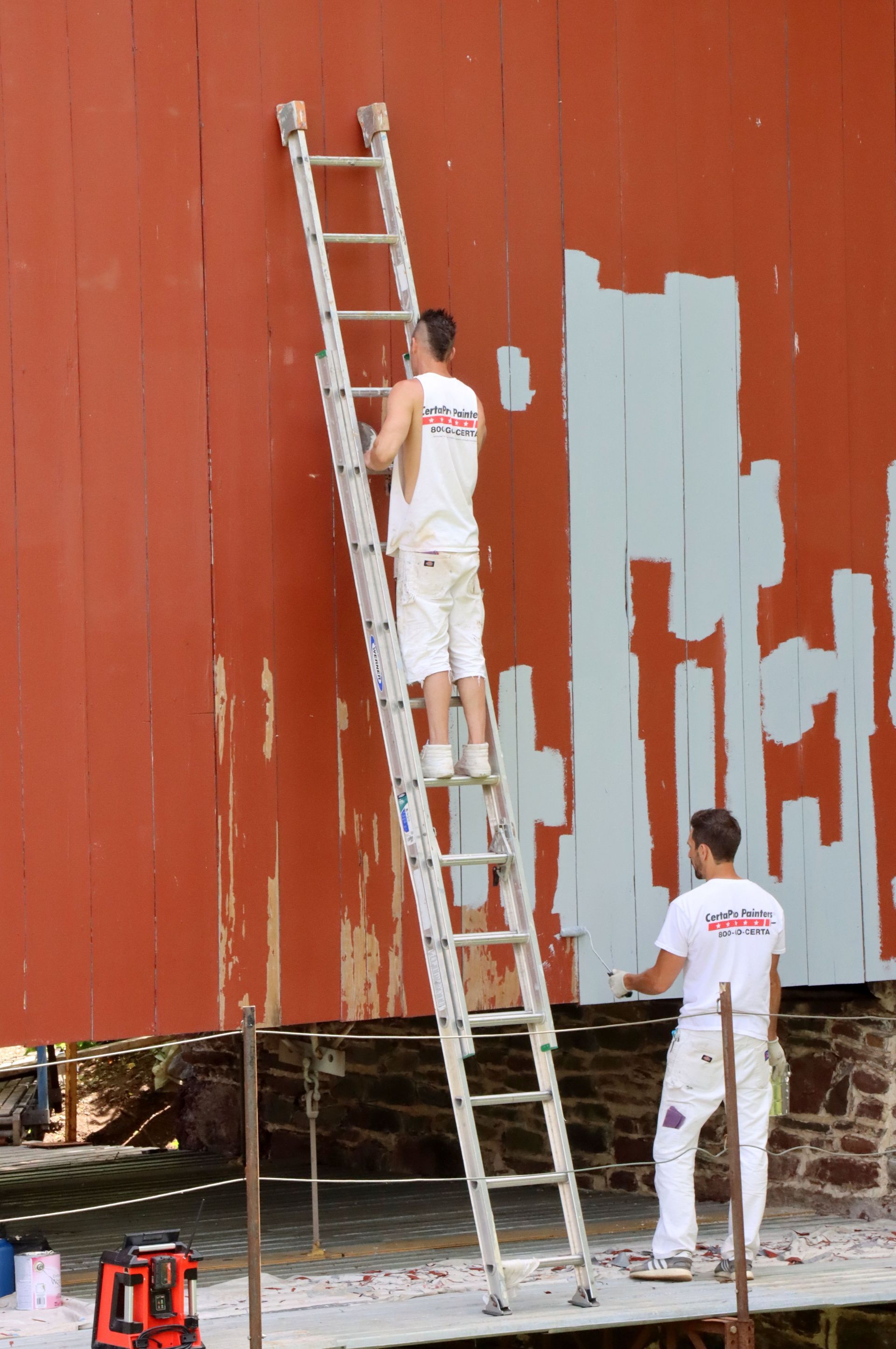 Two workers on ladders painting a red building wall, with a gray patch of unfinished paint.