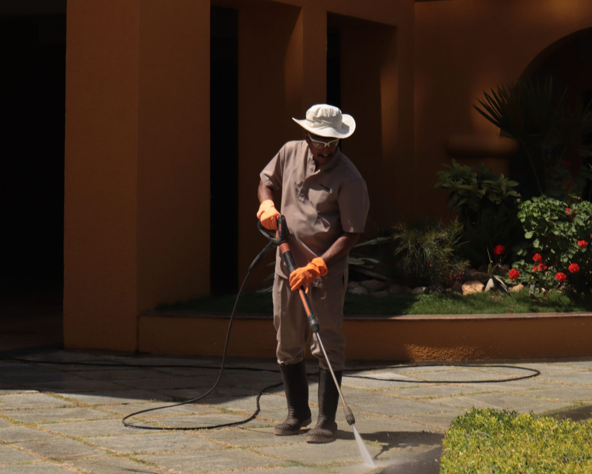 Worker with hose clearing the walkway outside a peach-colored building.