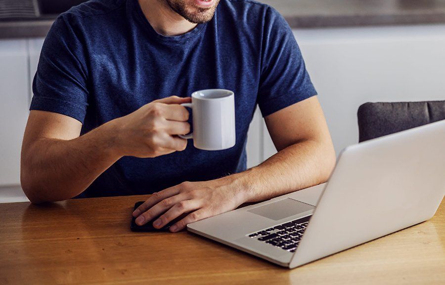 Un hombre está sentado en una mesa sosteniendo una taza de café y usando una computadora portátil.