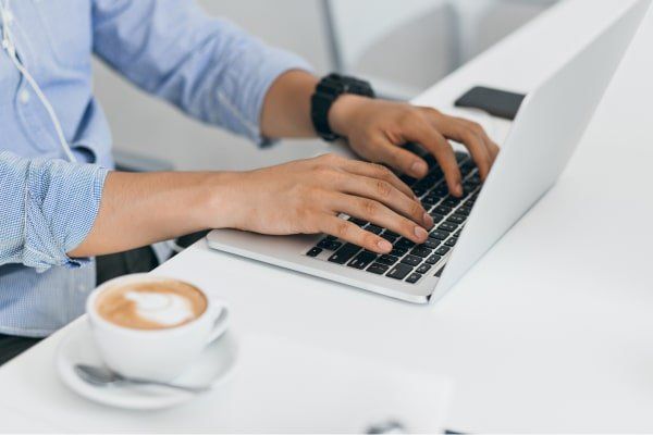 Una persona está escribiendo en una computadora portátil al lado de una taza de café.