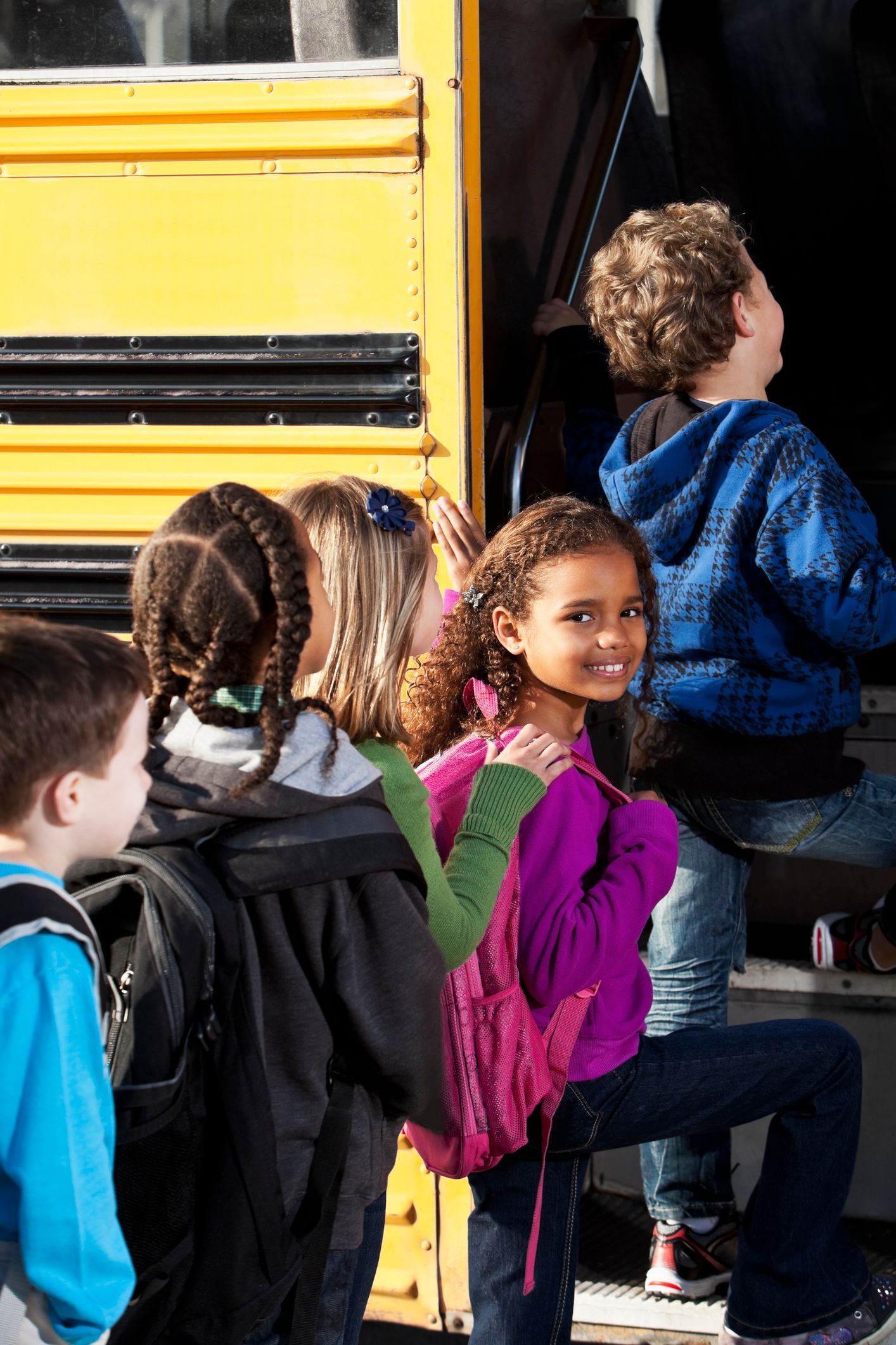 Children boarding a yellow school bus, one smiling at the camera, carrying backpacks. Children boarding a yellow school bus, one smiling at the camera, carrying backpacks.