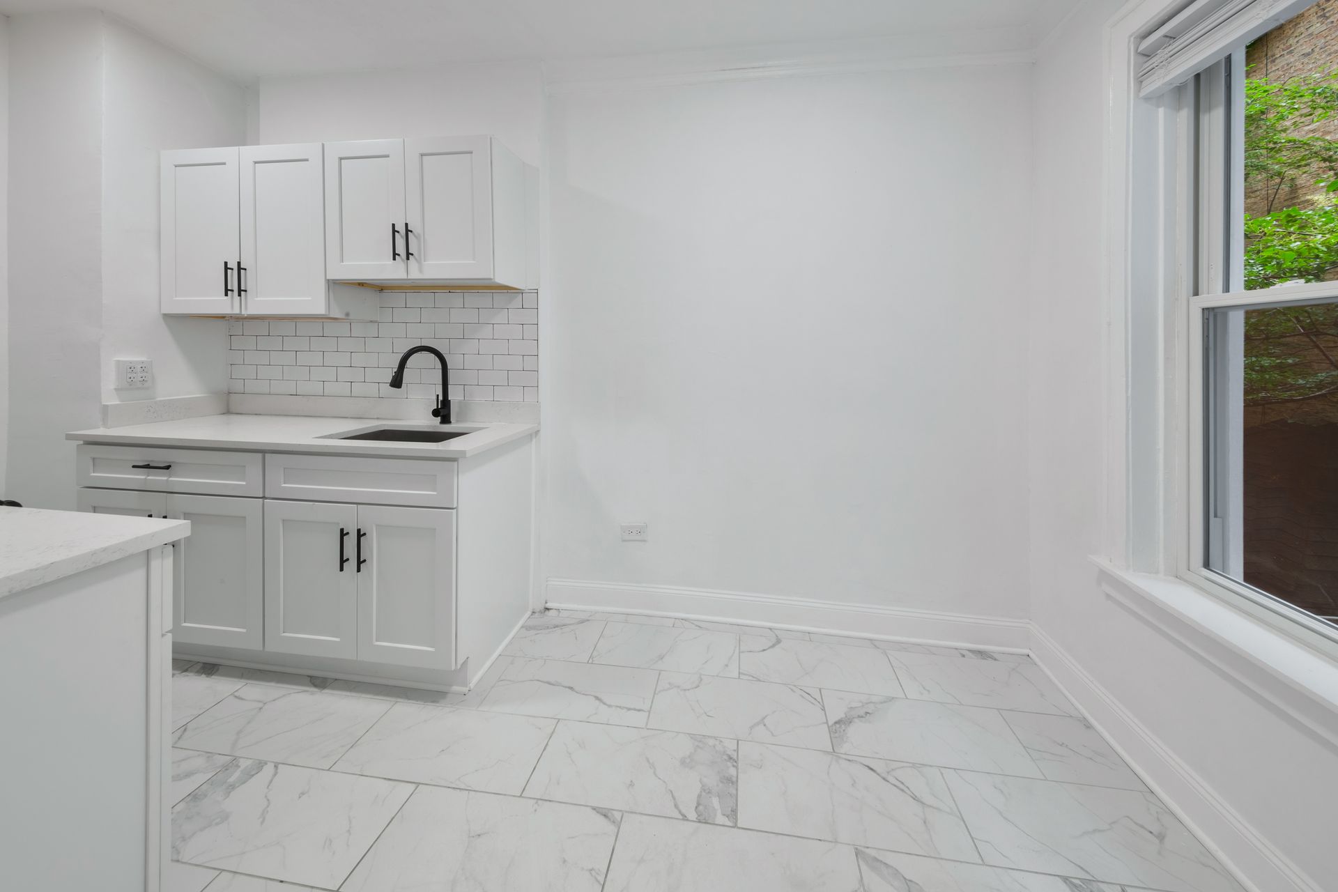 Small, all-white kitchen with white cabinets, sink, and marble-like floor tiles, and a window.