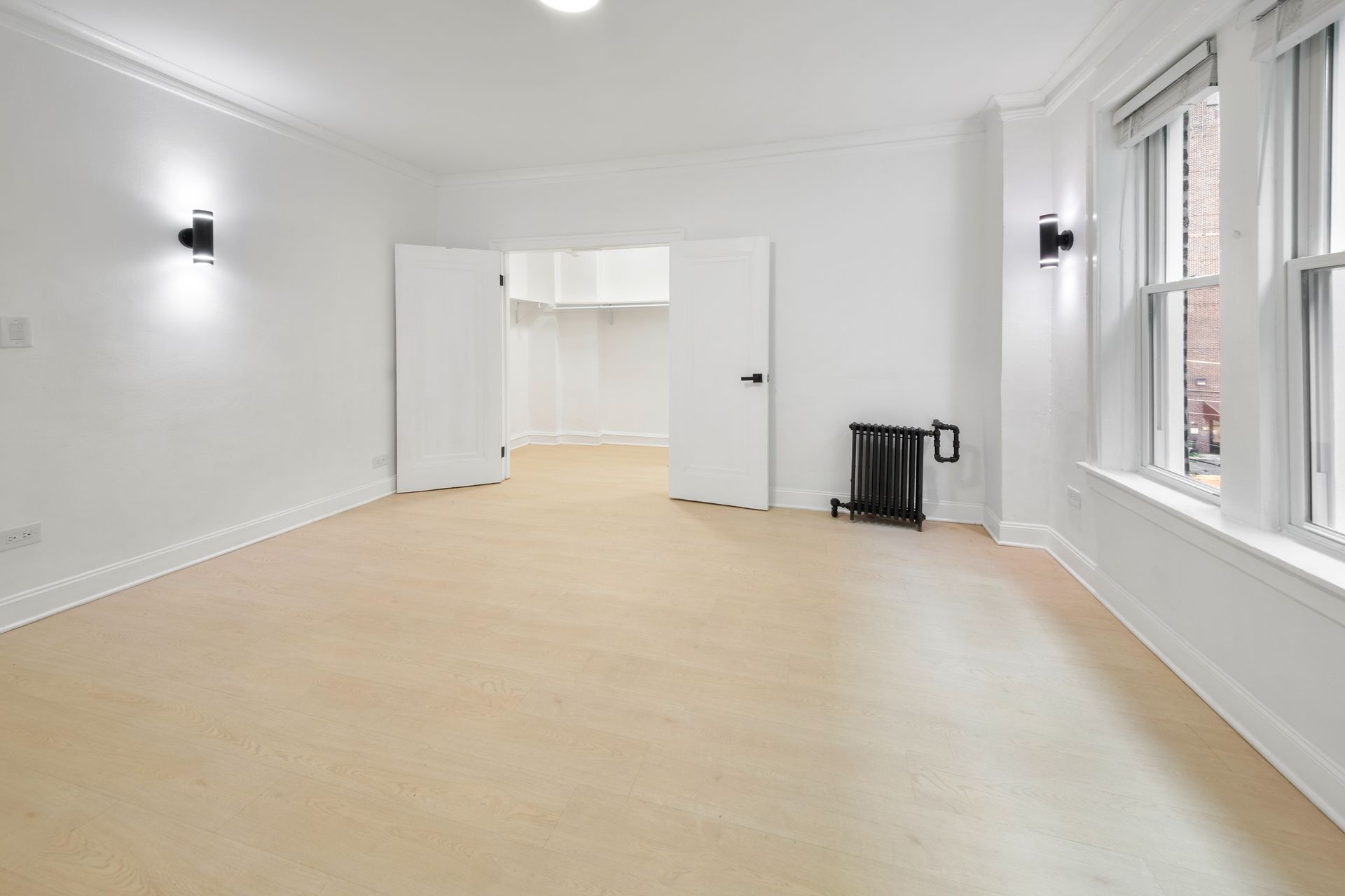 Empty white room with light-colored wood floors, two sconces, and a radiator.