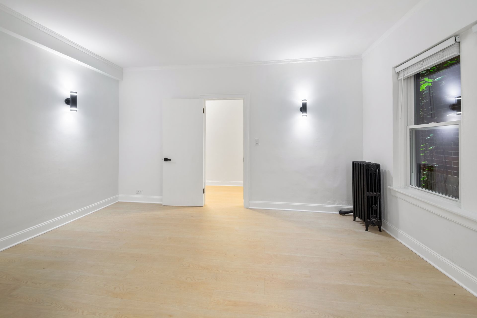 Empty, white-walled room with wood floor, two sconces, open door, radiator, and window.