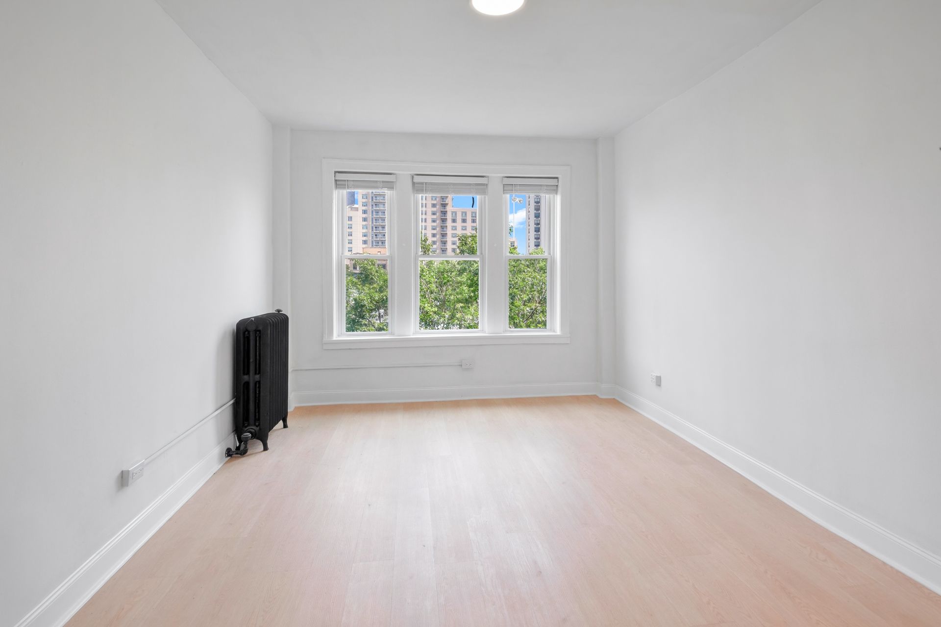 Empty room with white walls, three windows, radiator, and light wood floors.