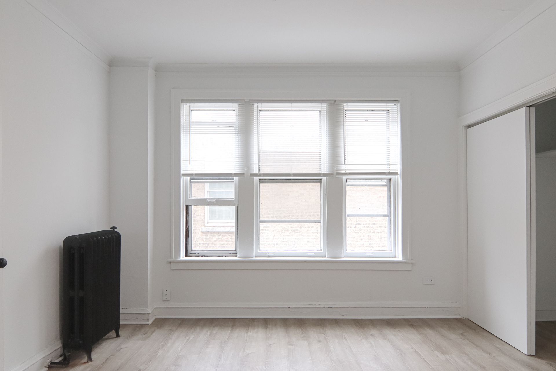Empty white room with a large window, closed blinds, and dark radiator. Closet on right.