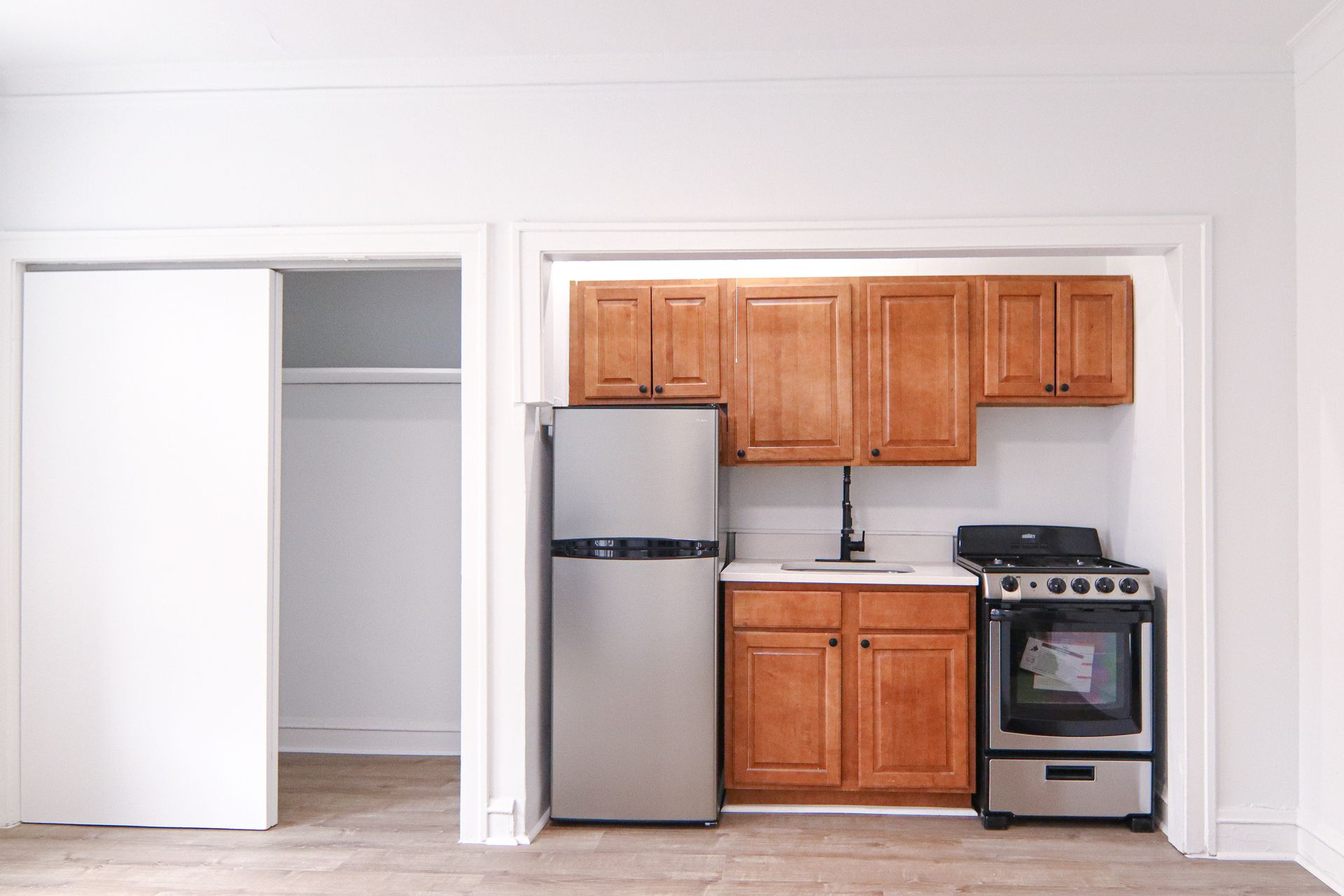 Small kitchen with wooden cabinets, stainless steel fridge, and gas stove in a white-walled room.