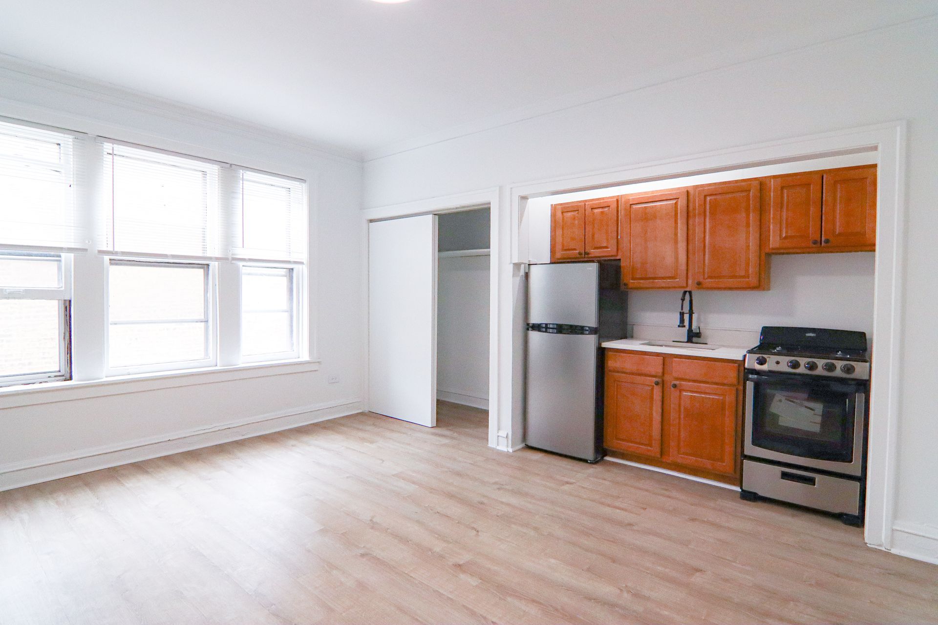An empty apartment with wood-look flooring, a small kitchen, and large windows.