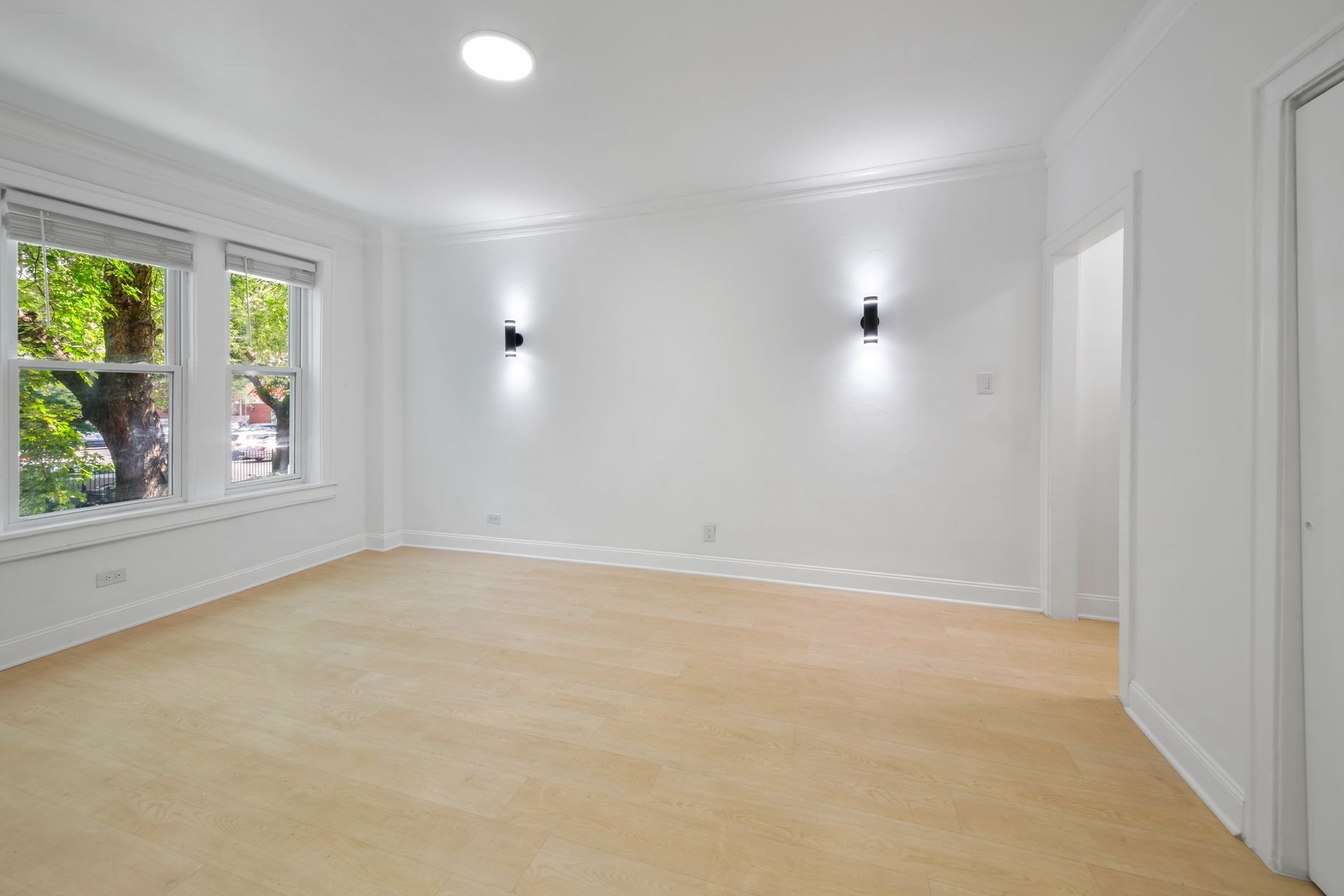 Empty, light-filled room with tan carpet, white walls, and two black wall sconces. Windows to the left.