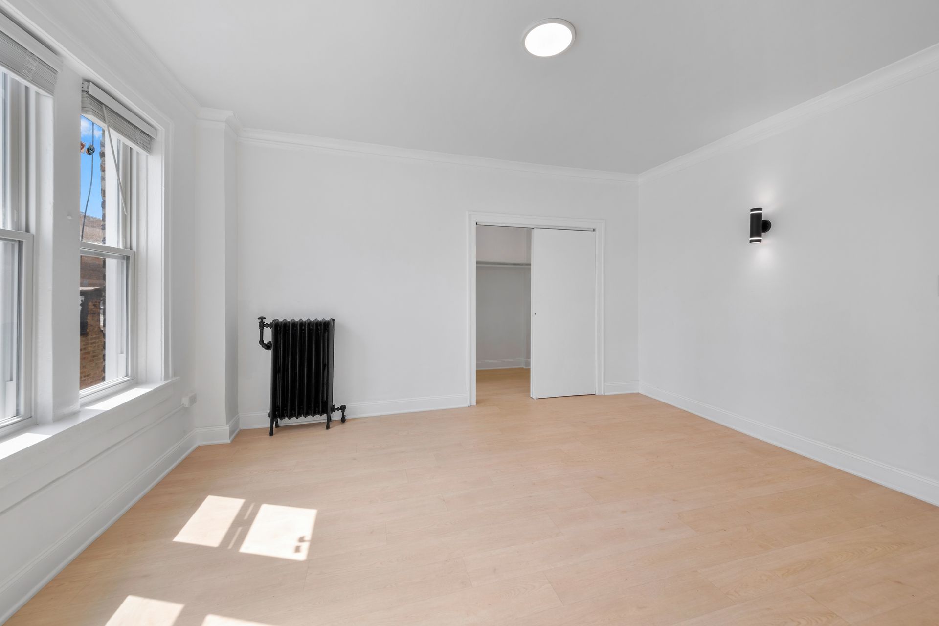 Empty white room with large window, beige carpet, black radiator, and closet door.