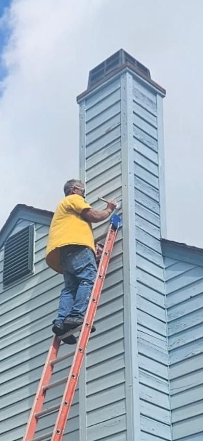 Man on ladder painting the exterior of a blue chimney. Yellow shirt and blue jeans are visible. Cloudy sky.