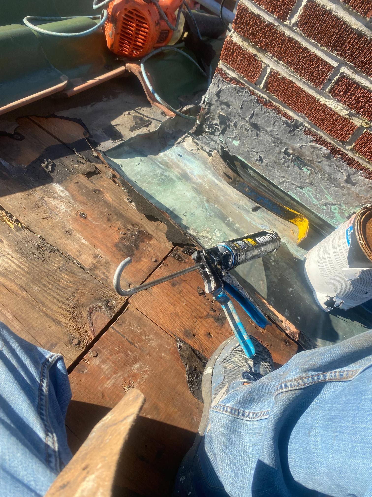 Person on a roof repairs copper flashing around a brick chimney, with tools and materials nearby.